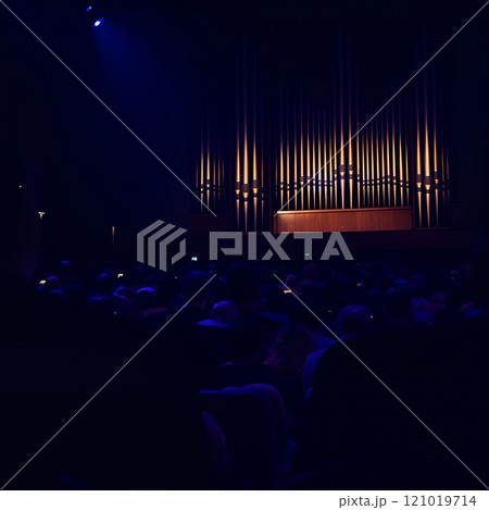 A large illuminated pipe organ in a dark concert hall with a seated audience, creating a dramatic and atmospheric scene 121019714