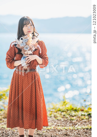 湖でドライフラワーを持って立っている赤いワンピースの女性 121023009