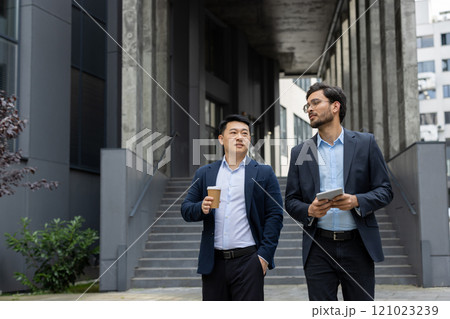 Two business professionals walk together outside an office building. One holds a coffee cup, while the other uses a tablet. They collaborate in a modern urban setting, fostering teamwork Two business professionals walk together outside an office building. One holds a coffee cup, while the other uses a tablet. They collaborate in a modern urban setting, fostering teamwork 121023239