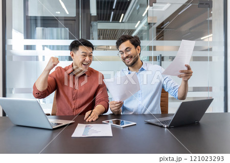 Business colleagues celebrate success with laptops and documents. Smiling coworkers review project results on paper and phone, highlighting teamwork and achievement in modern office. Business colleagues celebrate success with laptops and documents. Smiling coworkers review project results on paper and phone, highlighting teamwork and achievement in modern office. 121023293