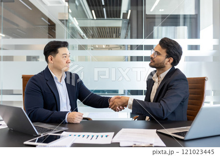 Businessmen shaking hands in modern office. Image captures partnership, trust, successful deal. Laptops, charts, documents on desk highlight professional environment. Concept of teamwork, cooperation 121023445