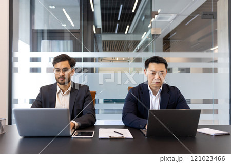 Two businessmen working on laptops in modern office. Collaborative professional setting conveys teamwork, productivity, focus. Ideal for business technology or professional workspace themes. 121023466