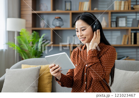 Asian woman at home enjoying music on headphones while using a tablet, seated on sofa in cozy living room. Scene depicts relaxation, technology, and casual leisure. 121023467