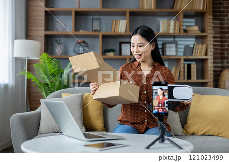 Asian woman creating video content at home, using phone to film while unboxing products. Surrounded by warm decor, she shares her excitement with viewers through engaging content. 121023499