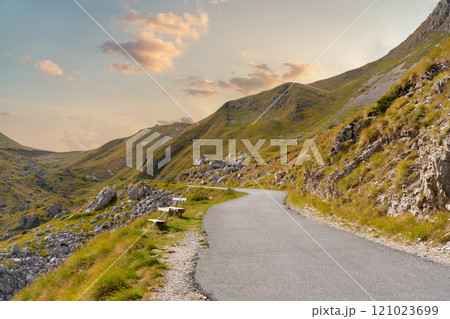 Winding mountain road surrounded by green slopes under a soft evening sky. 121023699