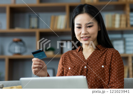 Smiling woman holds credit card while shopping online with laptop, enjoying convenience of e-commerce. Comfortable home setting reflects relaxed atmosphere and modern lifestyle. Smiling woman holds credit card while shopping online with laptop, enjoying convenience of e-commerce. Comfortable home setting reflects relaxed atmosphere and modern lifestyle. 121023941