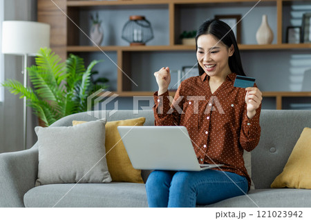 Asian woman celebrates online shopping success at home with laptop and credit card. Seated on couch, expressing excitement and joy, embodying satisfaction in modern e-commerce. Asian woman celebrates online shopping success at home with laptop and credit card. Seated on couch, expressing excitement and joy, embodying satisfaction in modern e-commerce. 121023942