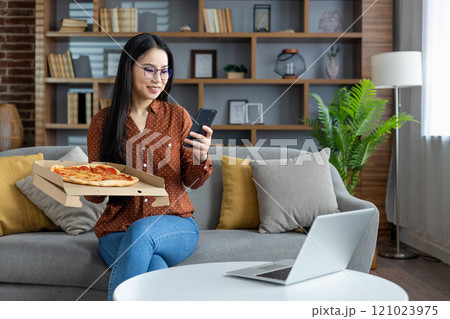 Asian woman enjoying pizza, using smartphone while sitting on sofa at home. Modern lifestyle embraces technology. Relaxed, casual moment with laptop, phone, and casual attire. Asian woman enjoying pizza, using smartphone while sitting on sofa at home. Modern lifestyle embraces technology. Relaxed, casual moment with laptop, phone, and casual attire. 121023975