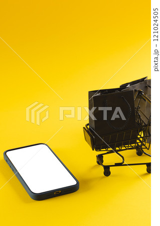 Vertical image of smartphone with copy space and trolley with bags on yellow background 121024505