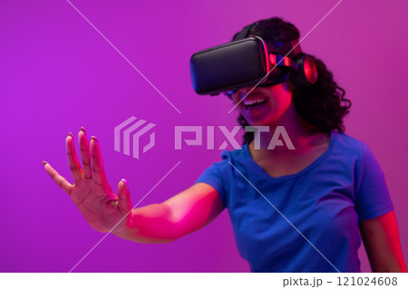 Biracial woman using vr headset and pointing on neon pink to purple background Biracial woman using vr headset and pointing on neon pink to purple background 121024608