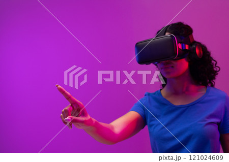 Biracial woman using vr headset and pointing on neon pink to purple background 121024609