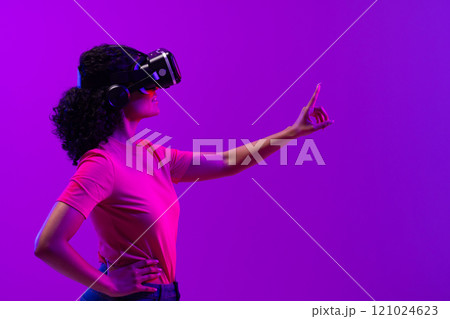 Biracial woman using vr headset and pointing on neon pink to purple background Biracial woman using vr headset and pointing on neon pink to purple background 121024623