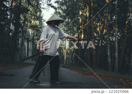 A martial artist in a conical Asian hat practices fighting techniques with a katana sword on a road in the forest. Katana training in the forest against the backdrop of autumn trees. A martial artist in a conical Asian hat practices fighting techniques with a katana sword on a road in the forest. Katana training in the forest against the backdrop of autumn trees. 121025066