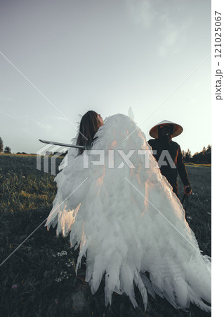 A samurai with a katana stabs an angel with large white wings in a dramatic scene set in a field at sunset, creating a powerful contrast between light and shadow. The struggle between good and evil. A samurai with a katana stabs an angel with large white wings in a dramatic scene set in a field at sunset, creating a powerful contrast between light and shadow. The struggle between good and evil. 121025067