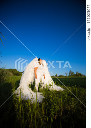 A woman with angel wings stands gracefully on a lush green field, looking up at the clear blue sky, creating a peaceful and magical scene. An angel against a blue sky looks up thoughtfully. 121025075