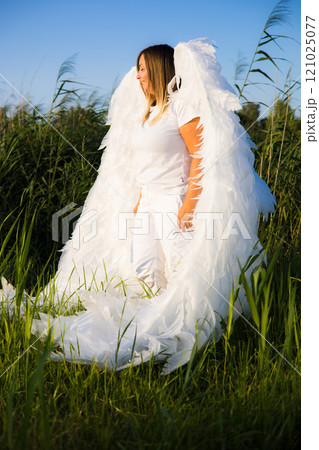 Blonde with big white angel wings standing on a field of tall grass against a blue sky enjoying the peaceful atmosphere. Cute girl as an angel in the rays of the setting sun. 121025077