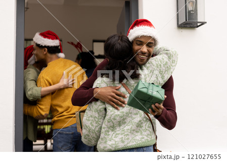 On doorstep, multiracial friends exchanging gifts and hugging at christmas party, wearing santa hats On doorstep, multiracial friends exchanging gifts and hugging at christmas party, wearing santa hats 121027655