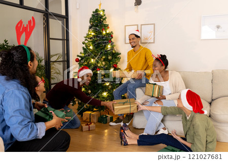 Multiracial friends exchanging gifts by christmas tree, wearing festive hats, at home Multiracial friends exchanging gifts by christmas tree, wearing festive hats, at home 121027681