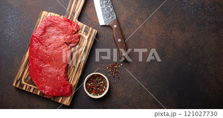 Raw uncooked top round beef steak on wooden cutting board with big kitchen knife and pepper on dark brown rustic stone background top view, cooking meat steak with copy space 121027730