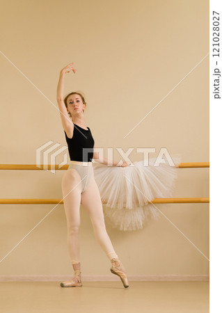 Ballet dancer gracefully raising arm while posing near barre Ballet dancer gracefully raising arm while posing near barre 121028027