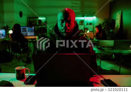Medium close up of African American brutal hacker programming laptop in room with other hackers in background 121028111