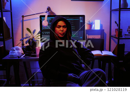 Portrait of female hacker dressed in black hoodie smiling lightly, table with computer, personal stuff and white mask behind her in dimly light room 121028119