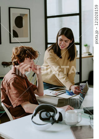 Vertical shot of young adult brunette female developer pointing at laptop screen while her coworker looking at it thoughtful 121028159