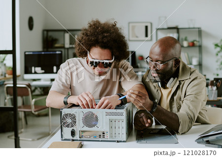 Medium close up of biracial colleagues repairing computer together at work desk, curly haired man connecting wires while his assistant holding blue wire 121028170