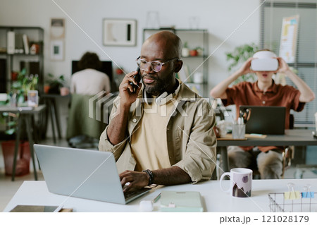 Medium close up of African American office worker working on laptop while speaking on smartphone 121028179