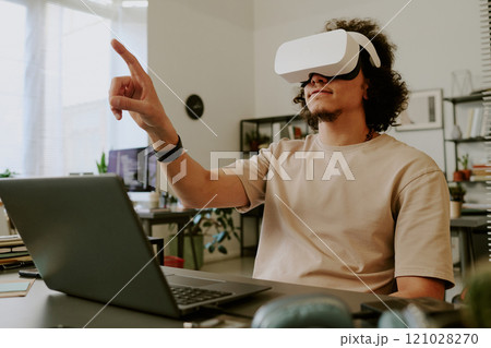 Low angle of young adult man sitting at desk with laptop and gesticulating while using VR goggles Low angle of young adult man sitting at desk with laptop and gesticulating while using VR goggles 121028270