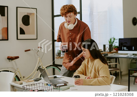 Caucasian young adult man with cup of hot coffee helping his female brunette colleague with laptop application while she sitting at work desk Caucasian young adult man with cup of hot coffee helping his female brunette colleague with laptop application while she sitting at work desk 121028308