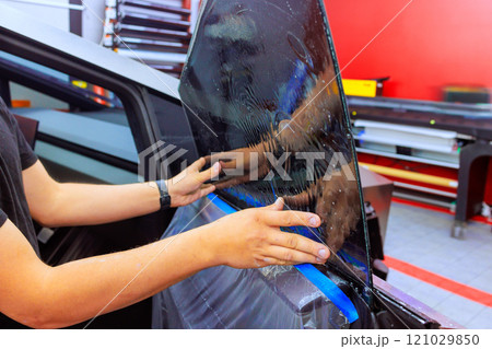 Technician worker applies tint film to car window in well lit automotive workspace, ensuring perfect fit. 121029850