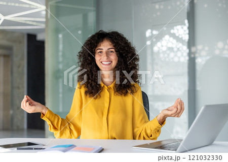 A smiling woman practices meditation at her office desk, conveying relaxation. The modern office environment enhances a sense of balance and peace during a work break. A smiling woman practices meditation at her office desk, conveying relaxation. The modern office environment enhances a sense of balance and peace during a work break. 121032330