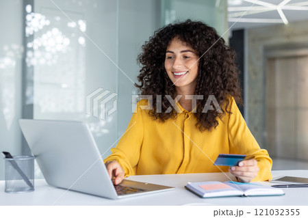 Smiling Latin American businesswoman using laptop and holding credit card. Engaged in online transaction or educational task. Represents modern business environment and online work. Smiling Latin American businesswoman using laptop and holding credit card. Engaged in online transaction or educational task. Represents modern business environment and online work. 121032355