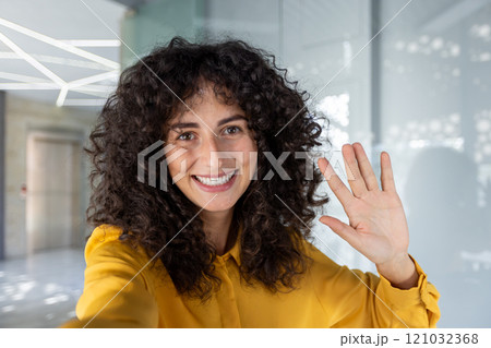 Woman with curly hair waves and smiles in office, capturing selfie with phone. Energy and friendliness radiate as she wears yellow shirt, set in modern business environment. Woman with curly hair waves and smiles in office, capturing selfie with phone. Energy and friendliness radiate as she wears yellow shirt, set in modern business environment. 121032368