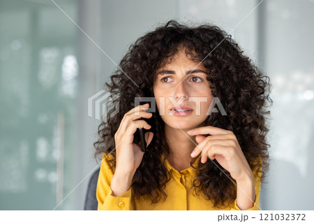 Curly-haired woman in yellow shirt using smartphone, appears concerned during phone call in office. Thoughtful and focused expression suggests work-related matters. Curly-haired woman in yellow shirt using smartphone, appears concerned during phone call in office. Thoughtful and focused expression suggests work-related matters. 121032372
