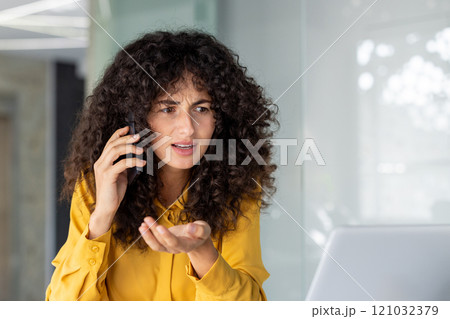 A woman talks on her phone, looking confused. She's in a modern office setting with a laptop, possibly dealing with a surprising situation. A woman talks on her phone, looking confused. She's in a modern office setting with a laptop, possibly dealing with a surprising situation. 121032379
