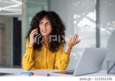 Confident young woman with engaged in animated phone conversation at desk. Laptop and documents on desk. Office environment suggests workplace dynamics. Confident young woman with engaged in animated phone conversation at desk. Laptop and documents on desk. Office environment suggests workplace dynamics. 121032380