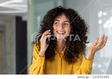 Joyful Latin American businesswoman with curly hair enjoying phone call. She exudes positivity while talking in contemporary office setting. Perfect for communication or business themes. 121032383