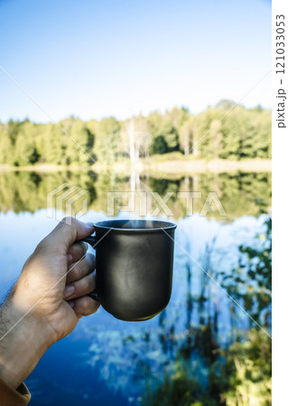 Hand holding a cup of drink in outdoor camp. Hot coffee, tea or mulled wine in a camp in nature in the forest. Travel, nature, recreation Hand holding a cup of drink in outdoor camp. Hot coffee, tea or mulled wine in a camp in nature in the forest. Travel, nature, recreation 121033053