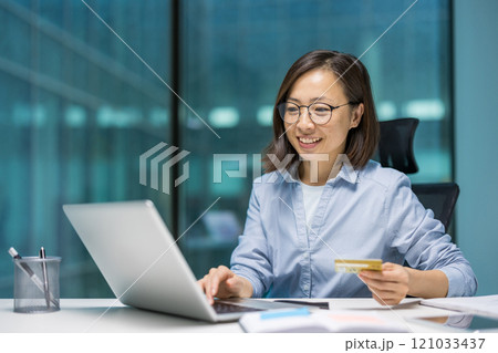 Asian businesswoman using laptop, holding credit card. Smiling, engaged in online shopping or electronic payment. Professional setting emphasizes modern business, finance, technology. Asian businesswoman using laptop, holding credit card. Smiling, engaged in online shopping or electronic payment. Professional setting emphasizes modern business, finance, technology. 121033437