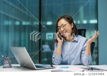 Asian businesswoman on phone call in office. Smiling and confident while using laptop, showcasing modern work environment and communication. Captures professionalism and productivity. Asian businesswoman on phone call in office. Smiling and confident while using laptop, showcasing modern work environment and communication. Captures professionalism and productivity. 121033464