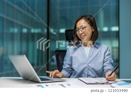 Asian businesswoman focused on laptop and documents at office desk. Professional setting emphasizing work, concentration, and efficiency. Woman smiling, multitasking with pen and technology. Asian businesswoman focused on laptop and documents at office desk. Professional setting emphasizing work, concentration, and efficiency. Woman smiling, multitasking with pen and technology. 121033490
