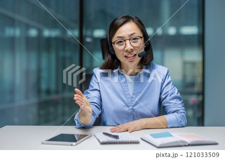 An Asian businesswoman using a headset to communicate during a virtual meeting. She is seated with documents and a tablet on the table. Ideal for concepts like communication An Asian businesswoman using a headset to communicate during a virtual meeting. She is seated with documents and a tablet on the table. Ideal for concepts like communication 121033509
