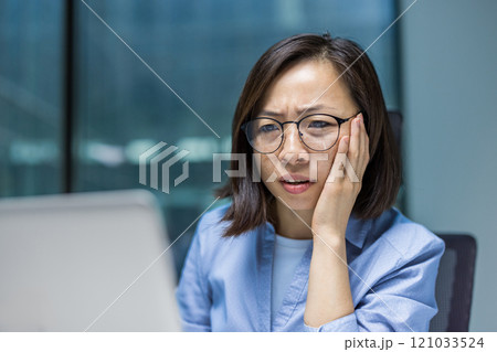 Asian businesswoman wearing glasses looks concerned while working on her laptop in an office setting. Her serious expression conveys focus and determination. Ideal for business-related content. 121033524