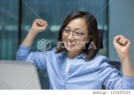 An Asian businesswoman joyfully raises her fists in celebration, highlighting her success at work. Her expression radiates happiness and achievement in a modern office 121033525