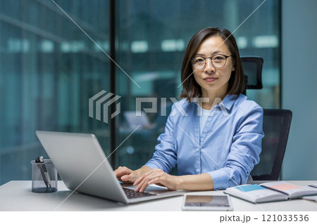Asian businesswoman focused on her laptop at a modern office desk. The image symbolizes productivity, concentration, and technology in a professional workplace environment. 121033536