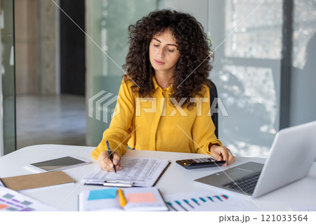Businesswoman in yellow blouse focused on calculations using documents and calculator at office desk with laptop and charts. Represents productivity, finance, and professional work setting. Businesswoman in yellow blouse focused on calculations using documents and calculator at office desk with laptop and charts. Represents productivity, finance, and professional work setting. 121033564