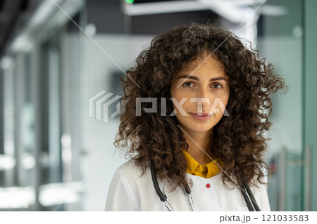 Professional female doctor with curly hair in hospital setting, wearing stethoscope. Represents healthcare, confidence, and medical service. Ideal image for medical and professional themes. 121033583