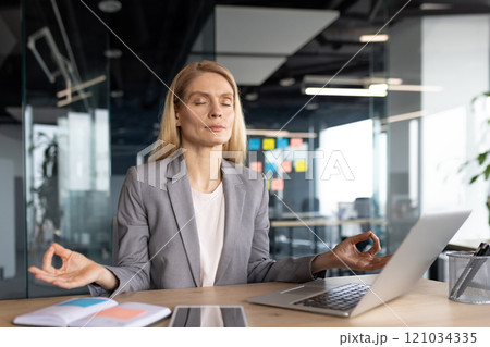 Mature woman businesswoman engaging in mindfulness meditation during office break. She sits calmly in front of laptop, seeking inner peace amidst workplace stress and daily challenges. 121034335